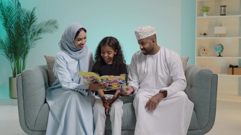 مفهوم التثقيف والبحث العلمي، هواية المطالعة والاستكشاف، قضاء وقت الفراغ بالتعلم والثقافة، بورتريه لطفلة عربية خليجية عمانية تجلس وتقوم بقراءة قصة لوالديها، اب يرتدي الزي التقليدي العماني، تنمية مهارات الطفل، شرح وتفسير أحداث القصة بأسلوب شيق وممتع، خلفية زرقاء