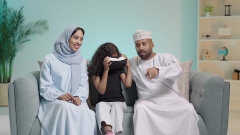 Spending enjoyable and entertaining times with virtual games, a portrait of an Arab Omani Gulf family, the girl wearing 3D glasses, enjoying watching virtual reality movies, simulating the fantasy world, a father wearing the traditional Omani dress, the concept of a happy family, blue background.