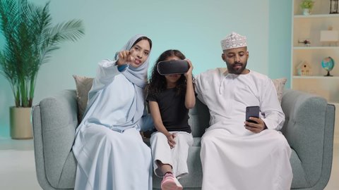 قضاء أوقات ممتعة ومسلية بالألعاب الافتراضية، بورتريه لعائلة عربية عمانية خليجية الطفلة ترتدي نظارة ثلاثية الأبعاد, الاستمتاع بمشاهدة أفلام الواقع الافتراضي، محاكاة العالم الخيالي، اب يرتدي الثوب العماني التقليديويحمل الهاتف المحمول، خلفية زرقاء