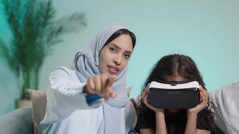 Spending enjoyable and entertaining times with virtual games, a close-up portrait of an Arab Omani Gulf girl wearing 3D glasses, enjoying watching virtual reality movies, simulating a fantasy world, a mother supporting and sharing playtime with her daughter wearing an abaya and hijab, blue background.