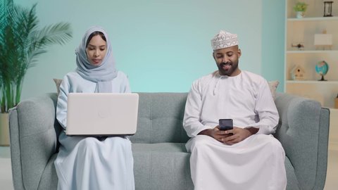 استخدام الأجهزة التقنية الحديثة, بورتريه لرجل عربي خليجي عماني يرتدي  الثوب ويجلس على آريكة ويحمل هاتف محمول بيده وبجابنبه زوجته تعمل على الحاسب المحمول، الدردشة مع الأهل والأصدقاء, تصفح برامج التواصل الاجتماعي، تطبيقات الأجهزة الحاسوبية، خلفية زرقاء