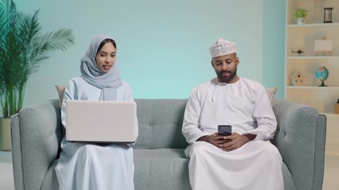 التحاور والنقاش في الرأي مع الأزواج، استخدام الأجهزة التقنية الحديثة, بورتريه لرجل عربي خليجي عماني يرتدي  الثوب ويجلس على آريكة ويحمل هاتف محمول بيده وبجابنبه زوجته تعمل على الحاسب المحمول، تصفح برامج التواصل الاجتماعي، تطبيقات الأجهزة الحاسوبية، خلفية زرقاء