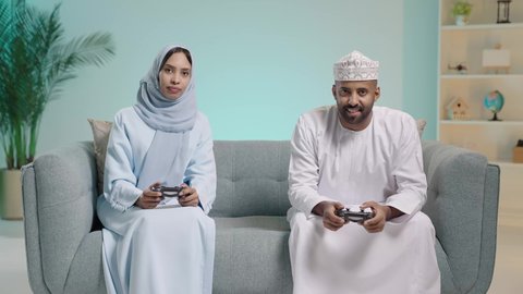 The concept of a happy family, enjoyment and entertainment with recreational PlayStation games, spending enjoyable time playing video games with gestures indicating happiness, a portrait of an Arab Gulf couple from Oman sitting in the living room holding a controller and playing video games competitively and cautiously, with a blue background.