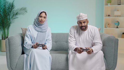 مفهوم المودة والتنافس معا، المتعة والتسلية  بألعاب البلايستيشن الترفيهية، قضاء وقت ممتع بلعب ألعاب الفيديو بإيماءات تدل على السعادة، بورتريه لزوجان عربيان خليجيان عمانيان يجلسان في غرفة المعيشة يمسكان عصا التحكم ويلعبان بألعاب الفيديو، خلفية زرقاء