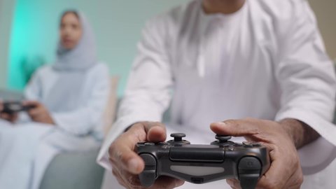 مفهوم المشاركة في التحديات والألعاب، المتعة والتسلية  بألعاب البلايستيشن الترفيهية، قضاء وقت ممتع بلعب ألعاب الفيديو بإيماءات تدل على السعادة، بورتريه مقرب لزوجان عربيان خليجيان عمانيان يجلسان في غرفة المعيشة يمسكان عصا التحكم ويلعبان بألعاب الفيديو، خلفية زرقاء