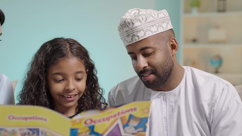 تنمية مهرات الطفل، هواية المطالعة والاستكشاف، قضاء وقت الفراغ بالتعلم والثقافة، مفهوم التثقيف والبحث العلمي، بورتريه لرجل عربي خليجي عماني يرتدي الثوب التقليدي يقوم بقراءة كتاب لطفلته، تنمية مهرات الطفل، شرح وتفسير أحداث القصة بأسلوب شيق وممتع، خلفية زرقاء