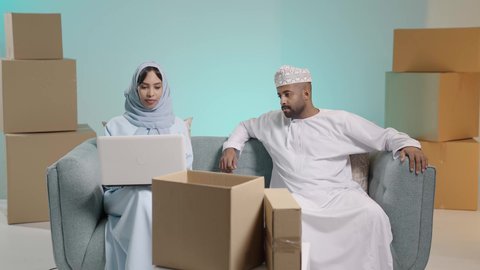 تطبيقات الأجهزة الحاسوبية، صناديق وكراتين، التحاور والنقاش في الرأي مع الأزواج، استخدام الأجهزة التقنية الحديثة, بورتريه لرجل عربي خليجي عماني يرتدي  الثوب ويجلس على آريكة وبجابنبه زوجته تعمل على الحاسب المحمول، تصفح برامج التواصل الاجتماعي، خلفية زرقاء