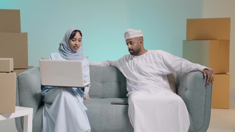 Boxes and cartons, computer applications, dialogue and discussion of opinions with spouses, use of modern technological devices, a portrait of an Arab Gulf Omani man wearing a dishdasha sitting on a sofa next to his wife who is working on a laptop, browsing social media, blue background.