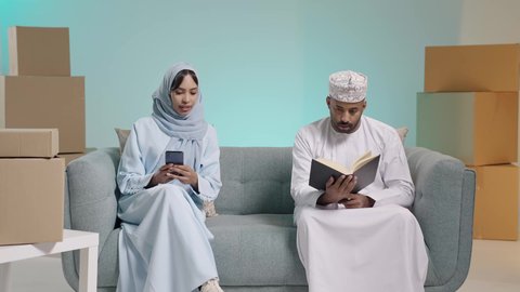 مفهوم المناقشة والحوار بين الأزواج، قضاء أوقات ممتعة في قراءة الكتب والقصص والروايات، بورتريه لرجل عربي خليجي عماني مثقف يرتدي الدشداشة والكمة يقوم بتصفح كتاب بجانبه زوجته تحمل الهاتف المحمول، مفهوم التعلم الذاتي والاطلاع والثقافة، خلفية زرقاء