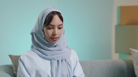 إنجاز مهام العمل عن بعد، ايماءات الاندماج والتركيز،التسوق الالكتروني وطلبات الاونلاين ، دردشة ومحادثات و تواصل اجتماعي ، اجراء اجتماع عمل عبر الانترنت، بورتريه مقرب لسيدة عربية عمانية خليجية تجلس على كرسي مريح وتستخدم الحاسوب المحمول، خلفية زرقاء