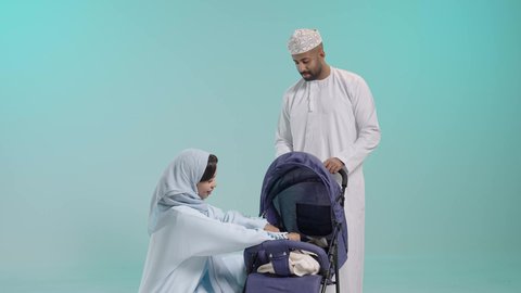 من الواجبات أساسية لدى الأهل متابعة تطور الطفل ونموه، القيام بنزهة وجولة عائلية، الرعاية والاهتمام بالأولاد, بورتريه لسيد عربي خليجي عماني يرتدي الثوب التقليدي يقف مع زوجته يقوم بجر عربة الطفل، القيام بنزهة وجولة عائلية، عائلة عمانية تقضي وقت ممتع، خلفية زرقاء