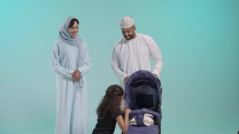 القيام بنزهة وجولة عائلية،أهمية الأهل ودورهم الأساسي في تربية الأبناء, الرعاية والاهتمام بالأولاد, بورتريه لسيد عربي خليجي عماني يرتدي الثوب التقليدي يقف مع زوجته وابنته يقوم بجر عربة الطفل، القيام بنزهة وجولة عائلية، عائلة عمانية تقضي وقت ممتع، خلفية زرقاء