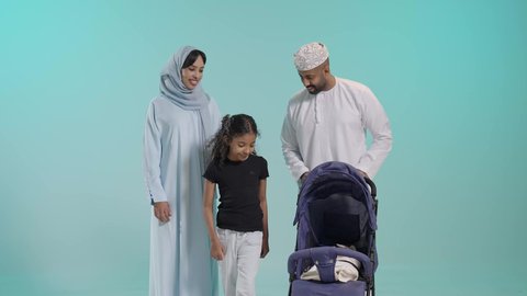 الرعاية والاهتمام بالأولاد, أهمية الأهل ودورهم الأساسي في تربية الأبناء, بورتريه لسيد عربي خليجي عماني يرتدي الثوب التقليدي يقف مع زوجته وابنته يقوم بجر عربة الطفل، القيام بنزهة وجولة عائلية، عائلة عمانية تقضي وقت ممتع، خلفية زرقاء