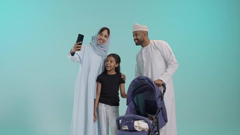 التقاط صورة سيلفي للمحافظة على الذكريات، القيام بنزهة وجولة عائلية، الرعاية والاهتمام بالأولاد, بورتريه لسيد عربي خليجي عماني يرتدي الثوب التقليدي يقف مع زوجته وابنته يقوم بجر عربة الطفل، القيام بنزهة وجولة عائلية، عائلة عمانية تقضي وقت ممتع، خلفية زرقاء