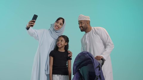 توثيق الذكريات من خلال الصور، القيام بنزهة وجولة عائلية، الرعاية والاهتمام بالأولاد, بورتريه لسيد عربي خليجي عماني يرتدي الثوب التقليدي يقف مع زوجته وابنته يقوم بجر عربة الطفل، القيام بنزهة وجولة عائلية، عائلة عمانية تقضي وقت ممتع، خلفية زرقاء