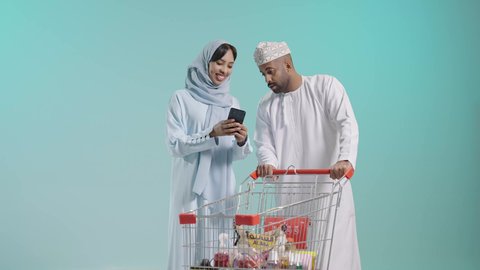 عربة تسوق تحتوي على منتجات غذائية ومنزلية، دمج التكنولوجيا في الحياة العملية، التعاون الأسري والاهتمام بشراء الأغذية الطازجة،الإستمتاع بالشراء والتبضع مع الأهل، بورتريه لزوجان عربيان خليجيان عمانيان يرتديان الزي التقليدي يقومان بالتسوق، خلفية زرقاء