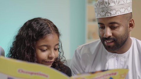 The hobby of reading and exploration, spending free time learning and gaining culture, the concept of education and scientific research, a portrait of an Arab Gulf Omani man wearing traditional attire sitting and reading a book to his daughter, developing the child's skills, explaining and interpreting the events of the story in an interesting and enjoyable way, blue background.