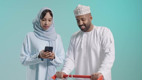 دمج التكنولوجيا في الحياة العملية، التعاون الأسري والاهتمام بشراء الأغذية الطازجة،الإستمتاع بالشراء والتبضع مع الأهل، بورتريه مقرب لزوجان عربيان خليجيان عمانيان يرتديان الزي التقليدي يقومان بالتسوق،عربة تسوق تحتوي على منتجات غذائية ومنزلية، خلفية زرقاء