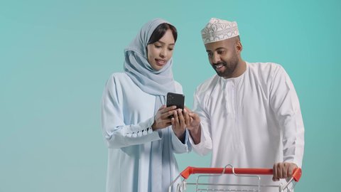 Various goods and food products, displayed against a green screen background, family cooperation and interest in buying fresh food, enjoying shopping and browsing with family, a portrait of an Arab Gulf couple from Oman wearing traditional attire while shopping, a shopping cart containing food and household products.