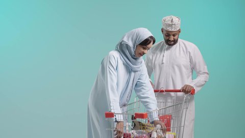 التعاون الأسري والاهتمام بشراء الأغذية الطازجة، السلع والمواد الغذائية المختلفة, الإستمتاع بالشراء والتبضع مع الأهل،زوجان عربيان خليجيان عمانيان يرتديان الزي التقليدي يقومان بالتسوق، منتجات غذائية وطنية ، عربة تسوق تحتوي على منتجات غذائية ومنزلية