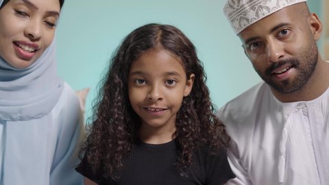 قضاء وقت ممتع بلعب ألعاب الفيديو بإيماءات تدل على السعادة، بورتريه مقرب لعائلة عربية خليجية عمانية تجلس في غرفة المعيشة، مفهوم الأسرة السعيدة، فتاه تمسك عصا التحكم وتلعب بألعاب الفيديو بجانب والديها ، المتعة و التسلية  بألعاب البلايستيشن الترفيهية، خلفية زرقاء