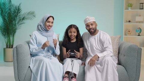 مفهوم الأسرة السعيدة، بورتريه لعائلة عربية خليجية عمانية تجلس في غرفة المعيشة ، قضاء وقت ممتع بلعب ألعاب الفيديو بإيماءات تدل على السعادة، فتاه تمسك عصا التحكم وتلعب بألعاب الفيديو بجانب والديها ، المتعة و التسلية  بألعاب البلايستيشن الترفيهية ، خلفية زرقاء