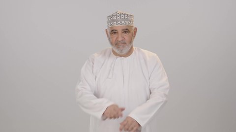 مفهوم الاهتمام بالمظهر الخارجي و الرجولة والأناقه، ايماءات الثقة بالنفس، بورتريه لرجل كبير بالسن عربي خليجي عماني يرتدي الدشداشة و الكمة يقف باستقامة بايدي متقاطعه, النظر إلى الكاميرا بتعابير السعادة والسرور، خلفية بيضاء
