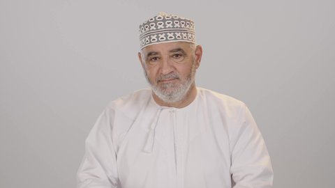 ايماءات الثقة بالنفس، مفهوم الاهتمام بالمظهر الخارجي و الرجولة والأناقه، بورتريه مقرب لرجل كبير بالسن عربي خليجي عماني يرتدي الدشداشة و الكمة يقف باستقامة بايدي متقاطعه, النظر إلى الكاميرا بتعابير السعادة والسرور، خلفية بيضاء