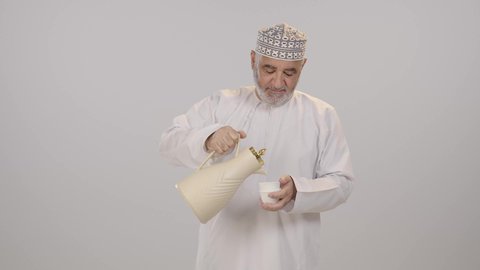 القيام بواجب استقبال الضيوف، ملامح الأصالة وروح التواصل الاجتماعي, بورتريه لرجل مسن عربي خليجي عماني مبتسم يرتدي الدشداشة و الكمة يحمل دلة تقليدية و يسكب القهوة في فنجان صغير، خلفية بيضاء