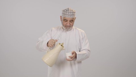 ملامح الأصالة وروح التواصل الاجتماعي, بورتريه لرجل مسن عربي خليجي عماني مبتسم يرتدي الدشداشة و الكمة يحمل دلة تقليدية و يسكب القهوة في فنجان صغير, تأدية واجب الضيافة، خلفية بيضاء