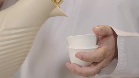 عادات استقبال الضيوف بروح الود والكرم, لقطة قريبة تبرز تفاصيل الضيافة العربية، بورترية مقرب ليد رجل عربي خليجي عماني يحمل دلة تقليدية و يسكب القهوة في فنجان صغير، خلفية بيضاء