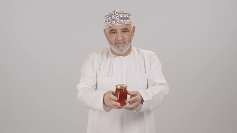 الأغذية الصحية المفيدة، منتجات غذائية محلية وطنية في سلطنة عمان، استخراج عسل النحل الطبيعي من المناحل، بورترية لرجل مسن عربي خليجي عماني يرتدي الدشداشة و الكمه يحمل بيده برطمان عسل نحلي ، خلفية بيضاء