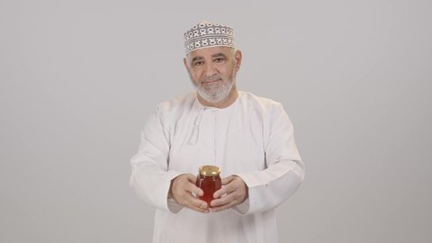 منتجات غذائية محلية وطنية في سلطنة عمان، استخراج عسل النحل الطبيعي من المناحل، الأغذية الصحية المفيدة، بورترية لرجل مسن عربي خليجي عماني يرتدي الدشداشة و الكمه يحمل بيده برطمان عسل نحلي ، خلفية بيضاء