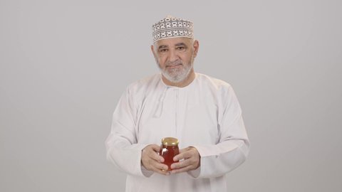 Extraction of natural honey from beehives, local national food products in the Sultanate of Oman, beneficial healthy foods, a portrait of an elderly Arab Gulf Omani man wearing a dishdasha and kumma holding a jar of honey in his hand, white background.