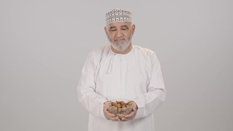 ابتسامة صادقة تعكس روح الكرم, الترحيب بالضيف وتقدير قيم المشاركة والود, بورتريه لرجل مسن عربي خليجي عماني يرتدي الدشداشة و الكمة يؤدي واجب الضيافة و يقدم صحن مليء بالتمر، خلفية بيضاء