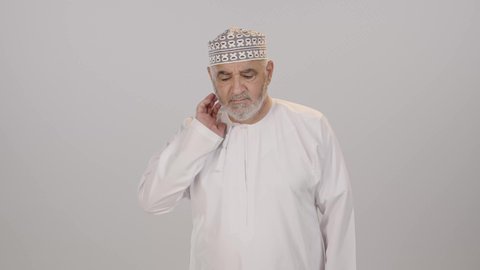التهاب العضلات والمفاصل، إيماءات الوجه تدل على الاضطراب والتألم، آلام الرقبة والعظام والعمود الفقري، ضرورة الذهاب للطبيب والحصول على العلاج، بورتريه لرجل مسن عربي خليجي عماني يرتدي الدشداشة و الكمه يمسك رقبته بتعابير الألم، خلفية بيضاء