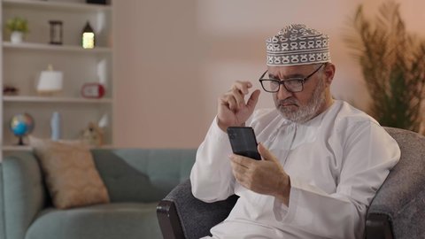 الدردشة والتواصل مع الأهل و الأصدقاء، ايماءات التركيز والجدية، مفهوم توضيح الصورة ، تصوير لرجل مسن عربي خليجي عماني يرتدي الدشداشة والكمة  و النظارة الطبية يجلس على الكرسي يحاول قراءة شي ما عبر الجوال