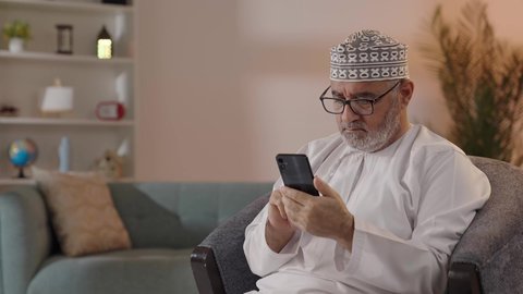 تصفح مواقع التواصل الاجتماعي، ايماءات التركيز والجدية، مفهوم توضيح الصورة ، تصوير لرجل مسن عربي خليجي عماني يرتدي الدشداشة والكمة  و النظارة الطبية يجلس على الكرسي يحاول قراءة شي ما عبر الجوال