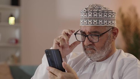 ايماءات التركيز والجدية، مفهوم توضيح الصورة ، تصويرمقرب  لرجل مسن عربي خليجي عماني يرتدي الدشداشة والكمة  و النظارة الطبية يحاول قراءة شي ما عبر الجوال ، الفحص والإستكشاف والقراءة