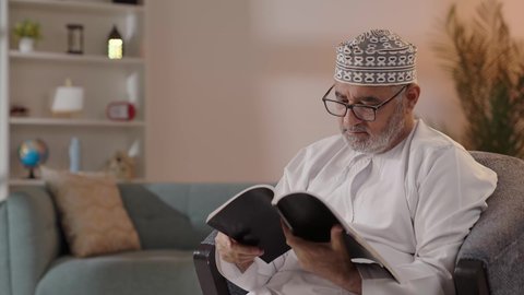 Utilizing free time for reading and knowledge, focusing on browsing useful books, the concept of contemplation and thinking, love for reading and literature, developing reading skills, a close-up image of an elderly Arab Gulf Omani man wearing a dishdasha and kumma and reading glasses sitting on a chair holding a book in his hand.