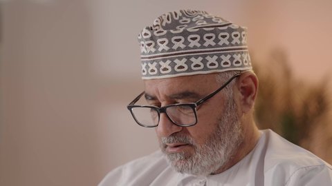 التعمق في الثقافة والاطلاع خلال وقت الفراغ، الاهتمام بتصفح الكتب المفيدة، مفهوم التأمل والتفكير, تنمية مهارات القراءة, تصوير مقرب لرجل مسن عربي خليجي عماني يرتدي الدشداشة والكمة و النظارة الطبية يمسك بيده الكتاب