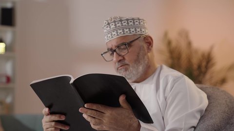 الاهتمام بتصفح الكتب المفيدة، مفهوم التأمل والتفكير، قضاء أوقات الفراغ بالتثقيف والتعلم، حب المطالعة والقراءة, تنمية مهارات القراءة, تصوير مقرب لرجل مسن عربي خليجي عماني يرتدي الدشداشة والكمة و النظارة الطبية يجلس على الكرسي ويمسك بيده الكتاب