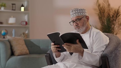 The concept of contemplation and thinking, spending free time on education and learning, love of reading and literature, developing reading skills, interest in browsing useful books, an elderly Arab Gulf Omani man wearing a dishdasha and a kumma and reading glasses sitting on a chair holding a book in his hand.