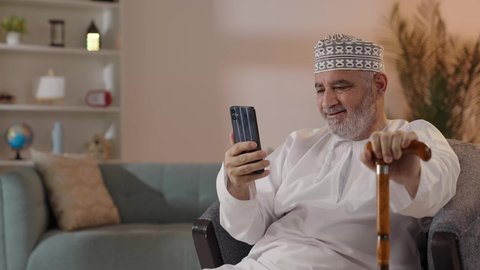 مكالمات فيديو للتواصل الاجتماعي عبر الجوال، ايماءات تدل على السعادة، استخدام التكنولوجيا و التقنيات الحديثة, تصويرمقرب لرجل مسن عربي خليجي عماني يرتدي الدشداشة و الكمة يجلس على الكرسي  يمسك  الهاتف المحمول و العكاز بيده