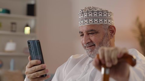 ايماءات تدل على السعادة، إجراء مكالمات فيديو مباشرة عبر الجوال، استخدام التكنولوجيا و التقنيات الحديثة, تصويرمقرب لرجل مسن عربي خليجي عماني يرتدي الدشداشة و الكمة يمسك  الهاتف المحمول و العكاز بيده