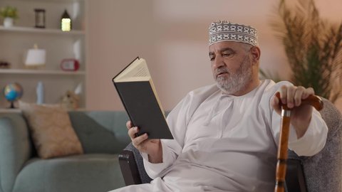 تنمية الذات خلال أوقات الفراغ بالاطلاع والتعلم، الاهتمام بتصفح الكتب المفيدة، مفهوم التأمل والتفكير، حب المطالعة والقراءة, تصوير لرجل مسن عربي خليجي عماني يرتدي الدشداشة والكمة  يجلس على الكرسي ويمسك بيده الكتاب و العكاز