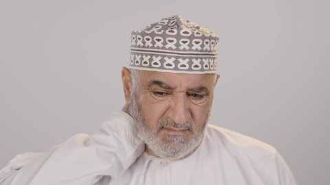 آلام الرقبة والعظام والعمود الفقري، إيماءات الوجه تدل على الاضطراب والتألم، ضرورة الذهاب للطبيب والحصول على العلاج، بورتريه مقرب لرجل مسن عربي خليجي عماني يرتدي الدشداشة و الكمه  يمسك رقبته بتعابير الألم، التهاب العضلات والمفاصل، خلفية بيضاء