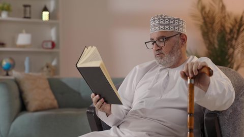 اكتساب المعرفة أثناء أوقات الفراغ، مفهوم التأمل والتفكير، حب المطالعة والقراءة, تنمية مهارات القراءة, الاهتمام بتصفح الكتب المفيدة, رجل مسن عربي خليجي عماني يرتدي الدشداشة والكمة و النظارة الطبية يجلس على الكرسي ويمسك بيده الكتاب و العكاز