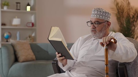 استغلال فترات الراحة في التعلم وتنمية الفكر, حب المطالعة والقراءة، رجل مسن عربي خليجي عماني يرتدي الدشداشة والكمة و النظارة الطبية يجلس على الكرسي ويمسك بيده الكتاب و العكاز