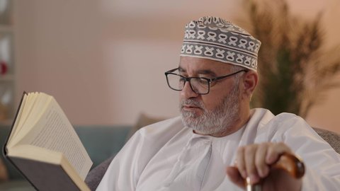 الاهتمام بالقراءة والتثقيف في أوقات الفراغ ، مفهوم التأمل والتفكير, تصوير مقرب لرجل مسن عربي خليجي عماني يرتدي الدشداشة والكمة و النظارة الطبية يجلس على الكرسي ويمسك بيده الكتاب و العكاز